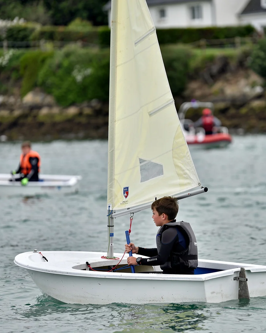 A young child aged around 6 to 7 years old navigating an Optimist dinghy on the calm, protected waters of étang de Dahouët in Pléneuf-Val-André during a spring